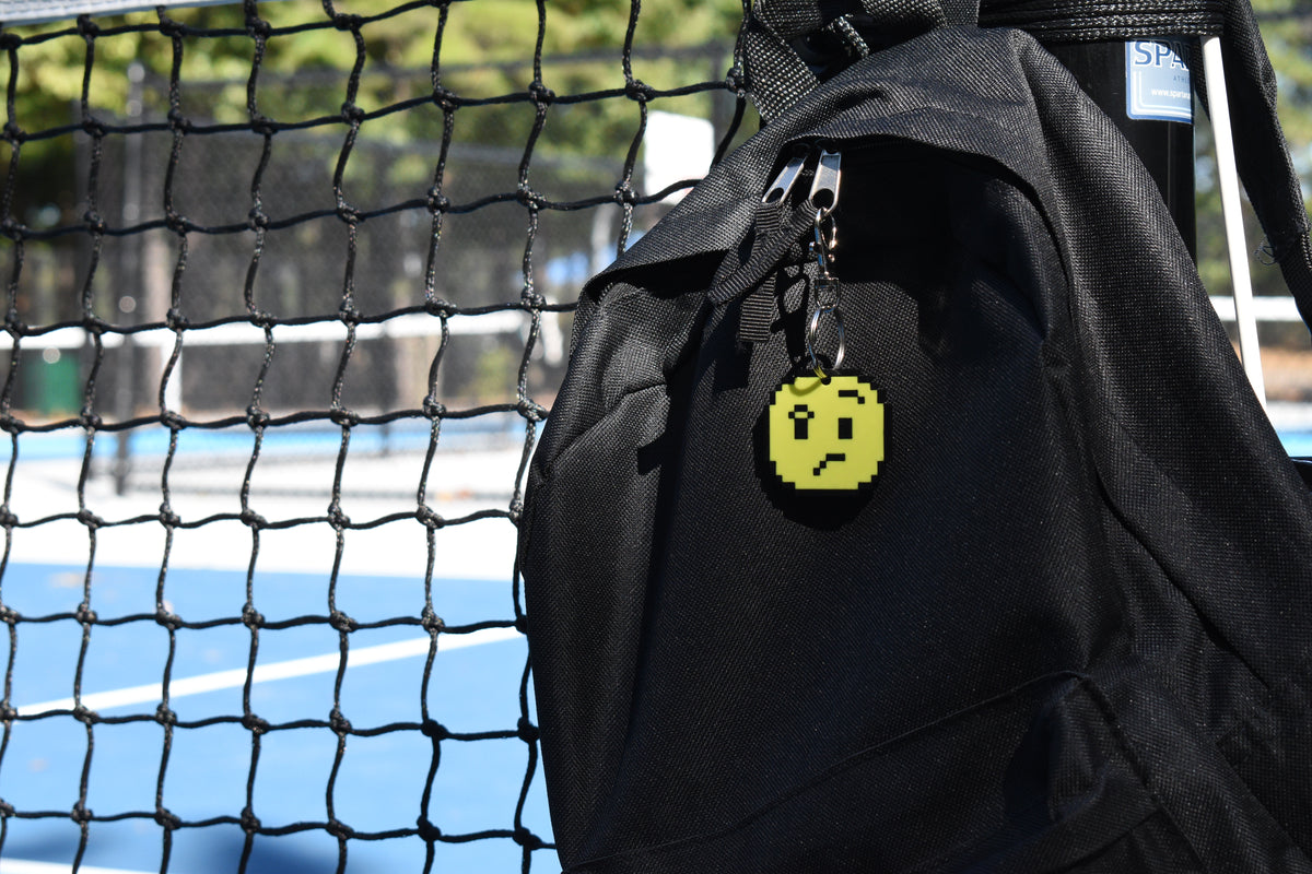 Pixelated confused face emoji keychain clipped to a black backpack zipper outdoors near a tennis court by LayerPlay