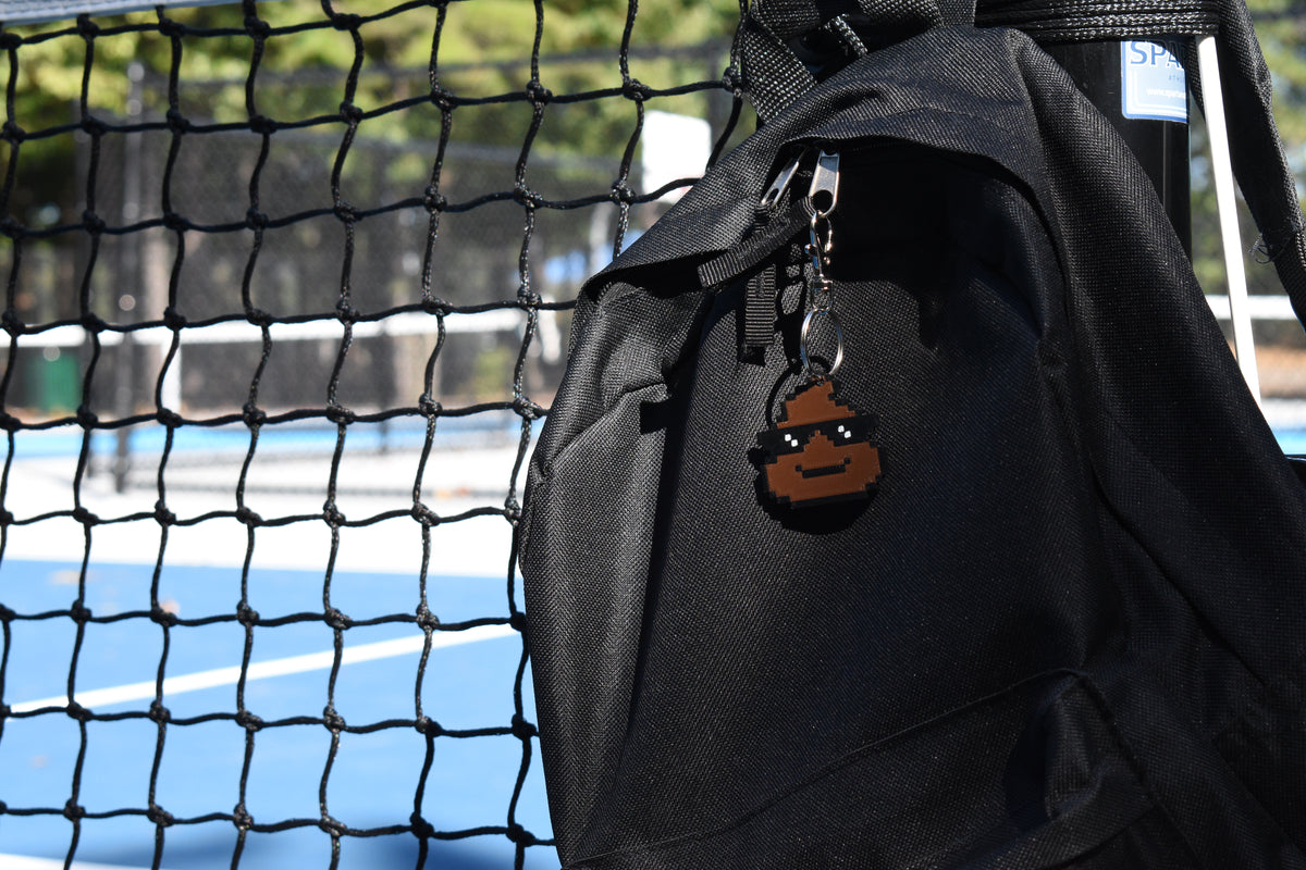 Pixelated cool poop emoji keychain clipped to a black backpack zipper outdoors near a tennis court by LayerPlay