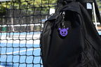 Pixelated devil emoji keychain clipped to a black backpack zipper outdoors near a tennis court by LayerPlay