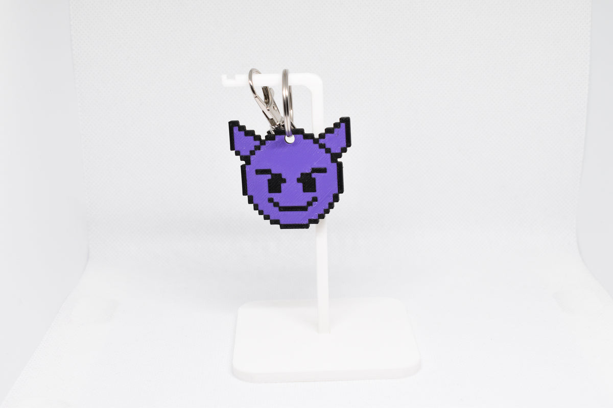 3D printed retro pixelated devil emoji keychain hanging on a white display stand by LayerPlay