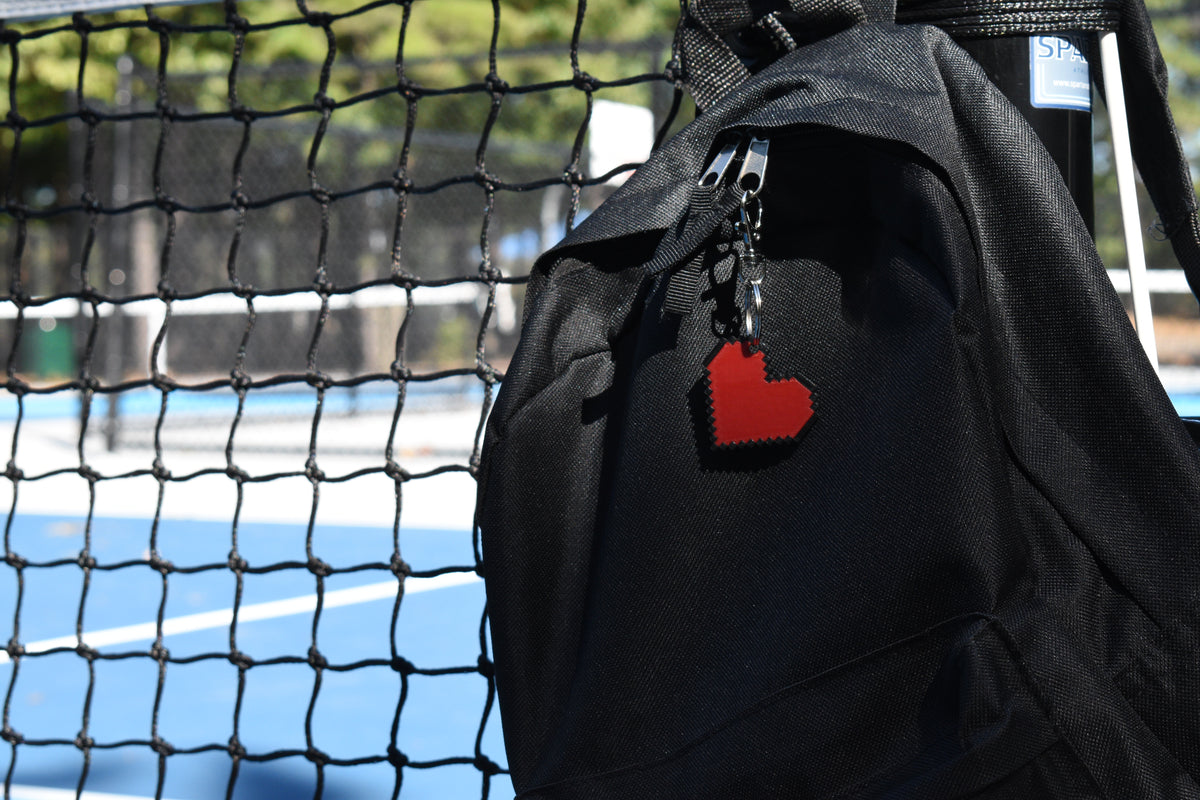 Pixelated heart emoji keychain clipped to a black backpack zipper outdoors near a tennis court by LayerPlay
