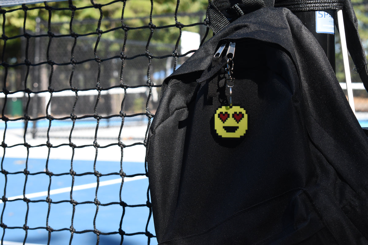 Pixelated heart eyes emoji keychain clipped to a black backpack zipper outdoors near a tennis court by LayerPlay