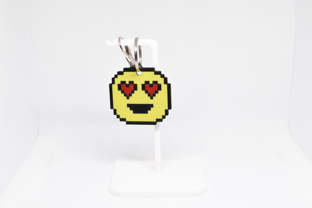 3D printed retro pixelated heart eyes emoji keychain hanging on a white display stand by LayerPlay