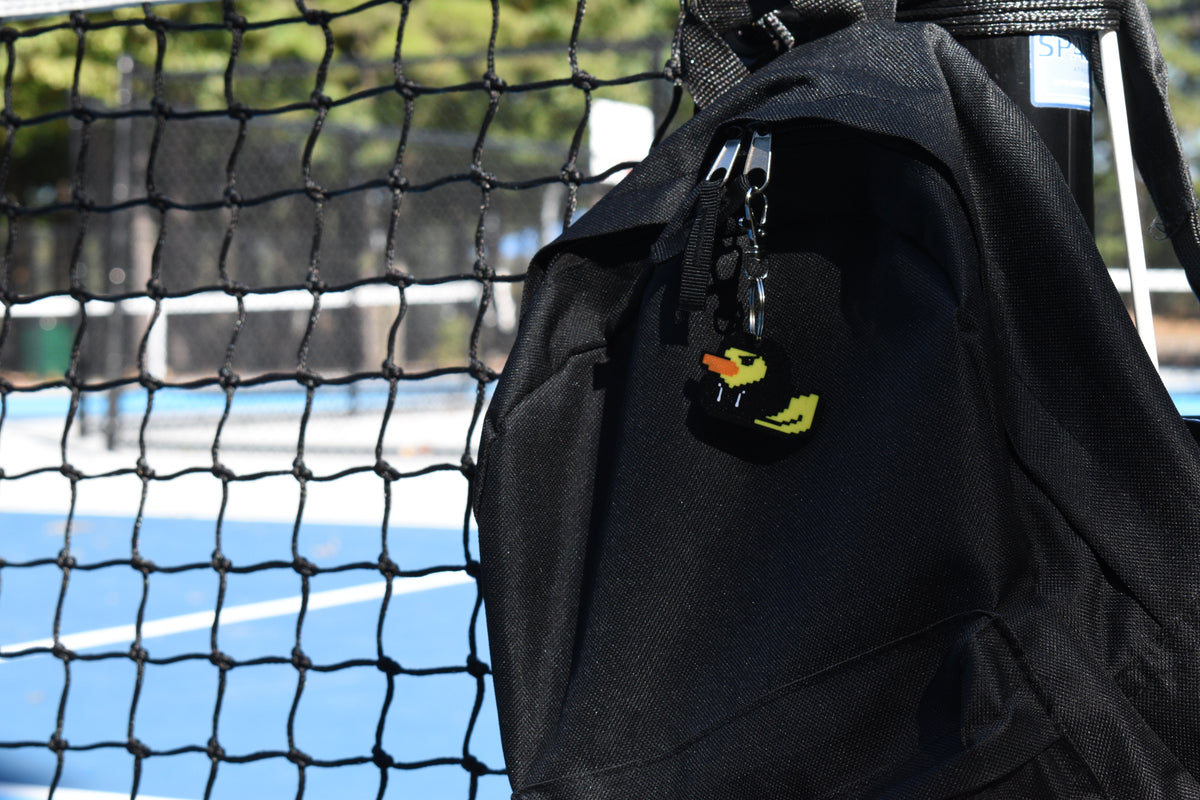 Hoodie Duck pixel rubber duck keychain clipped to a black backpack zipper outdoors near a tennis court by LayerPlay