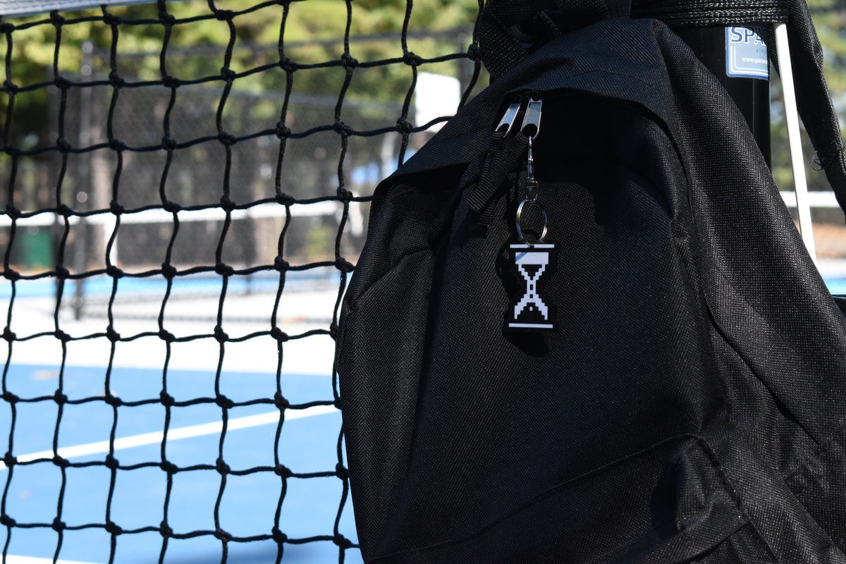Pixelated hourglass cursor keychain clipped to a black backpack zipper outdoors near a tennis court by LayerPlay