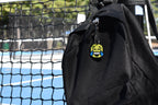 Pixelated laughing face emoji keychain clipped to a black backpack zipper outdoors near a tennis court by LayerPlay