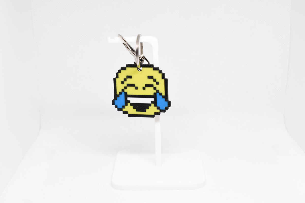 3D printed retro pixelated laughing face emoji keychain hanging on a white display stand by LayerPlay