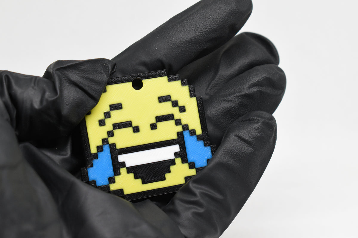 Hand in black glove holding 3D printed pixel laughing face emoji keychain front view detail by LayerPlay