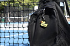 Lil Quack pixel rubber duck keychain clipped to a black backpack zipper outdoors near a tennis court by LayerPlay