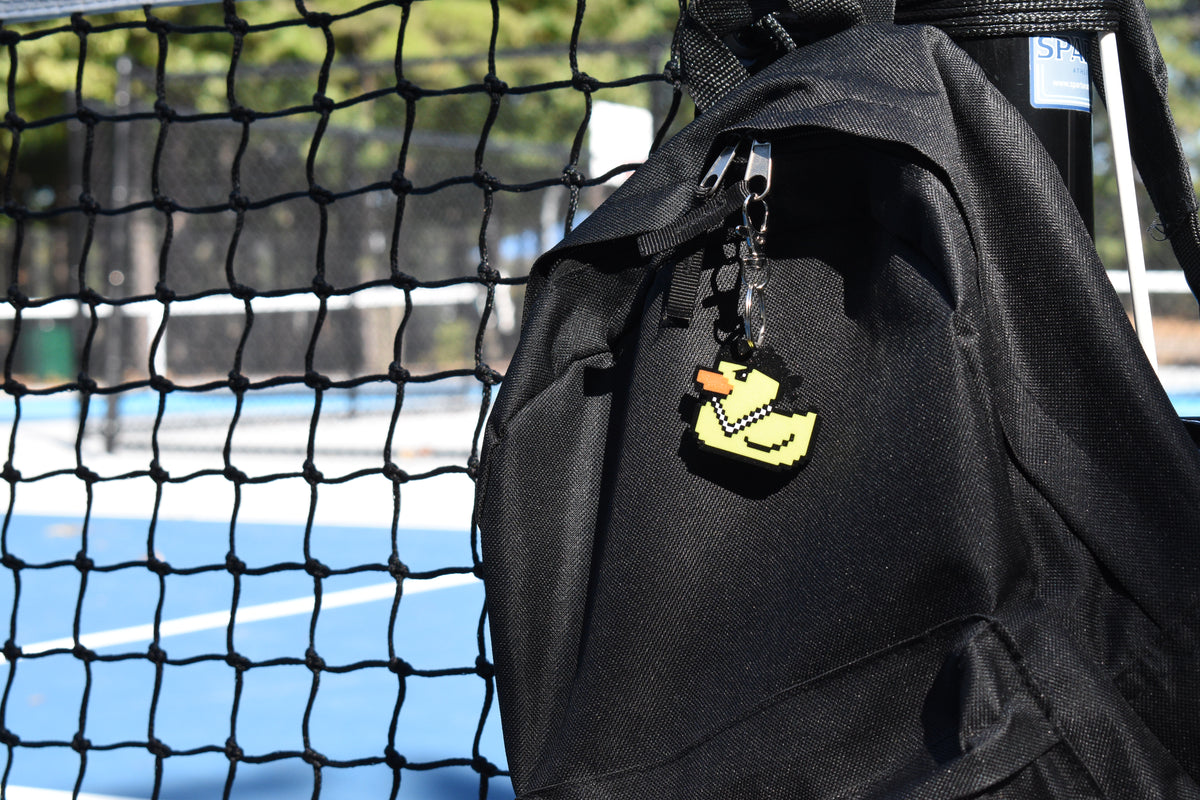 Lil Quack pixel rubber duck keychain clipped to a black backpack zipper outdoors near a tennis court by LayerPlay