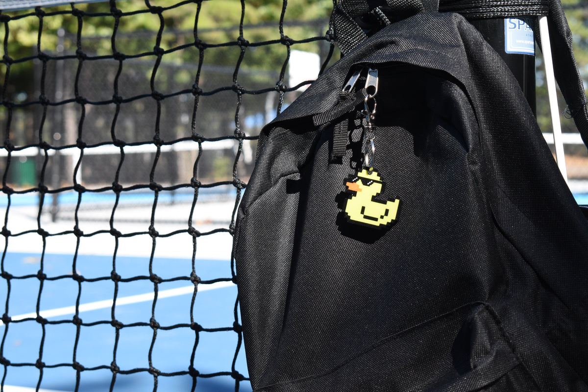 Pixel rubber duck keychain clipped to a black backpack zipper outdoors near a tennis court by LayerPlay