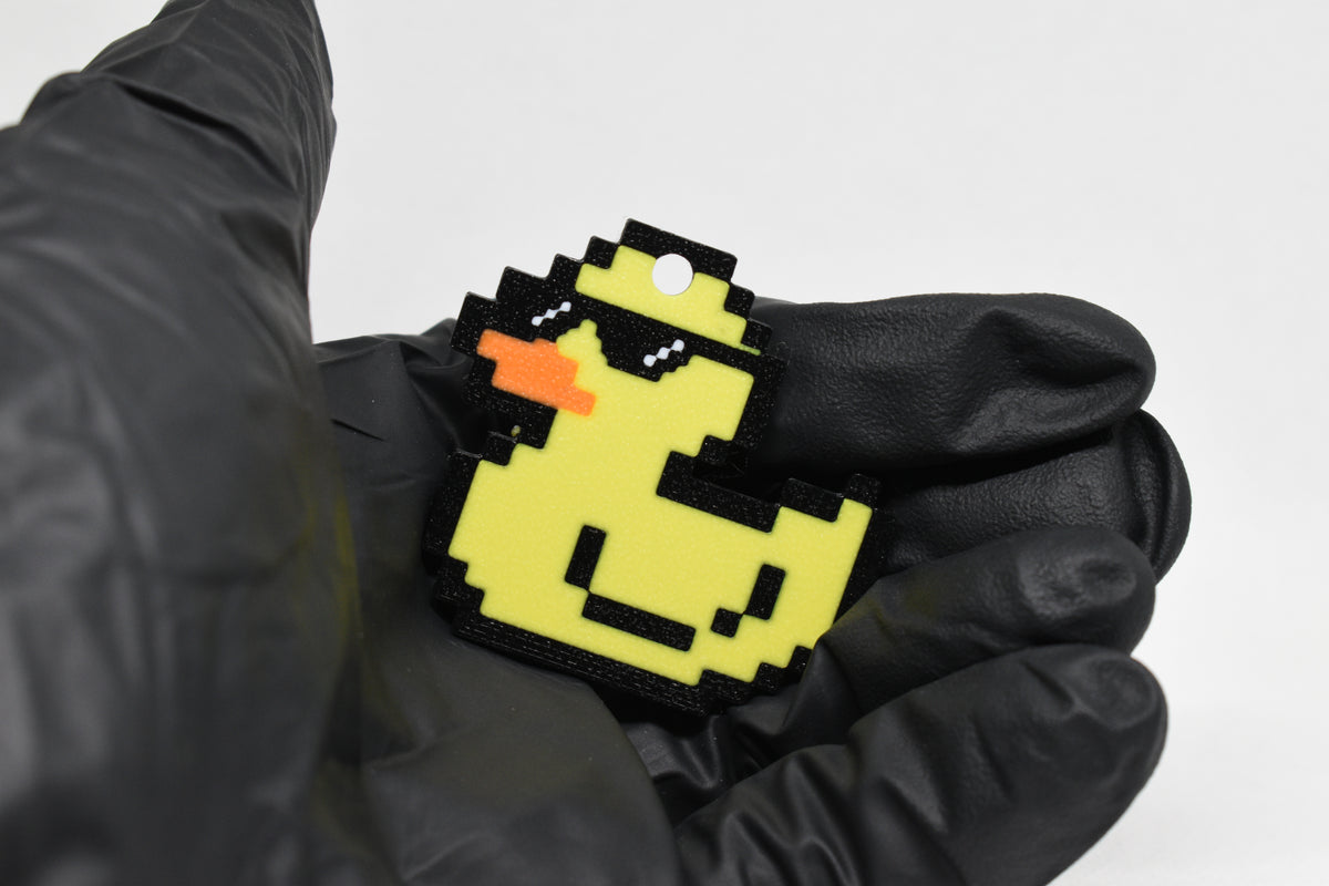 Hand in black glove holding 3D printed pixel rubber duck wearing sunglasses keychain front view detail by LayerPlay