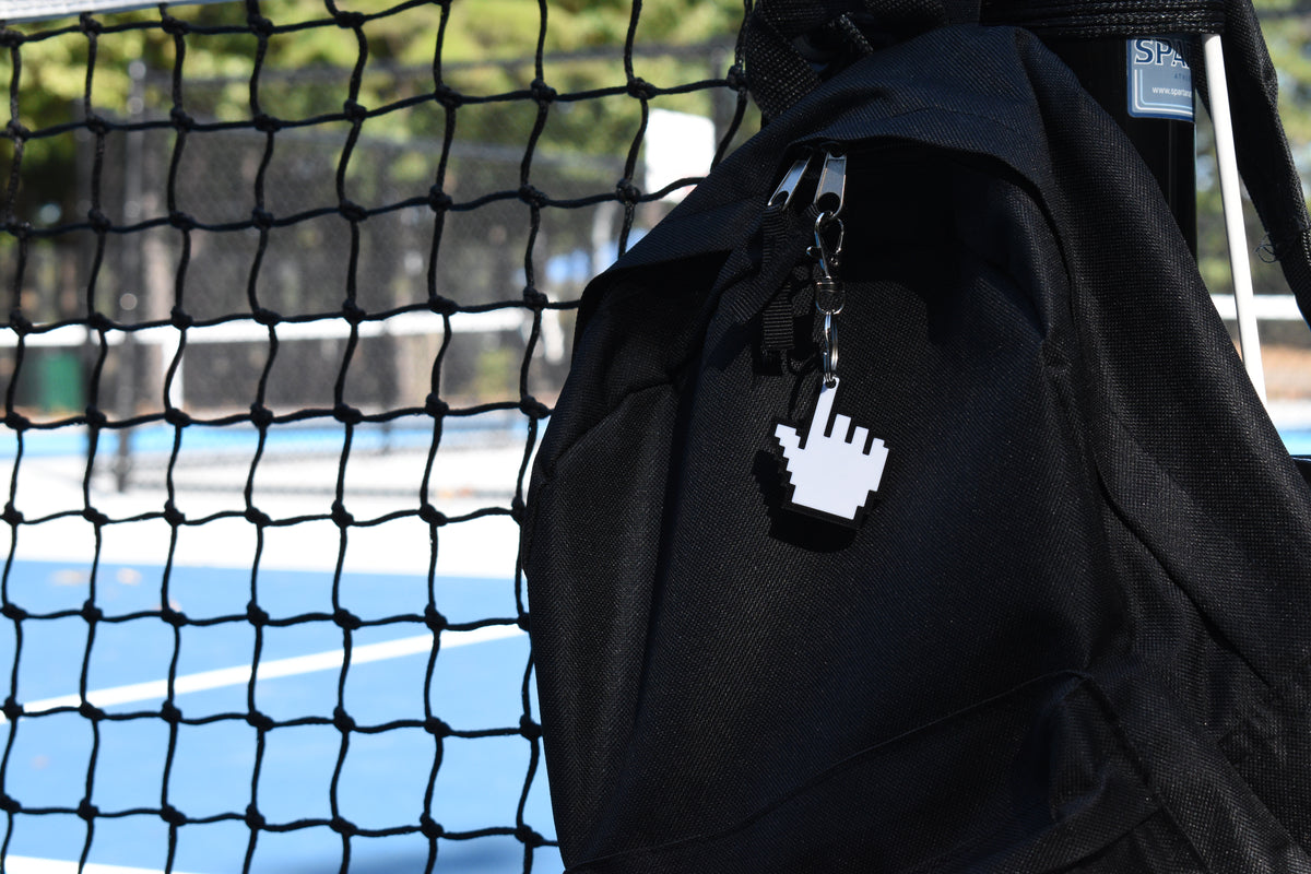 Pixelated pointing hand cursor keychain clipped to a black backpack zipper outdoors near a tennis court by LayerPlay