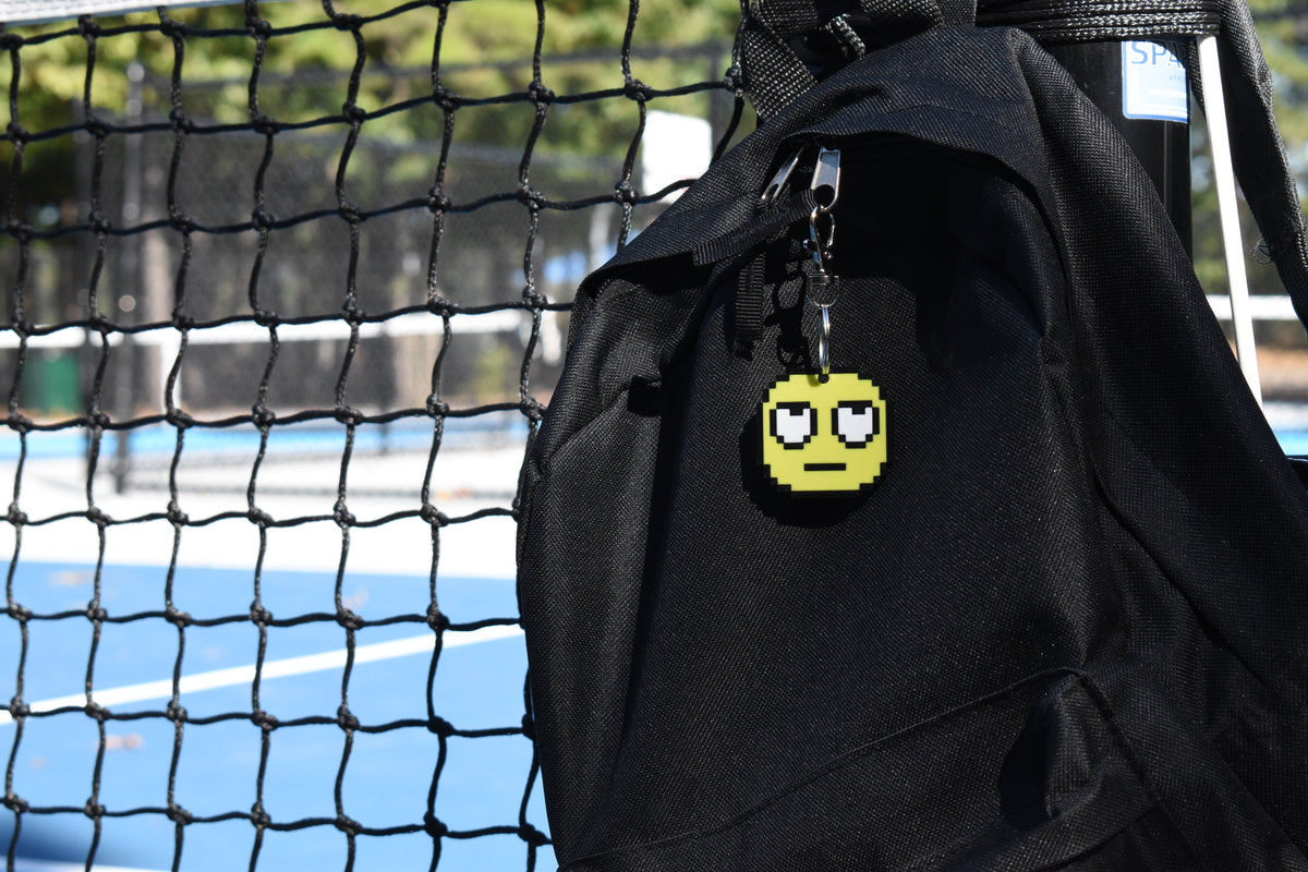 Pixelated rolling eyes emoji keychain clipped to a black backpack zipper outdoors near a tennis court by LayerPlay