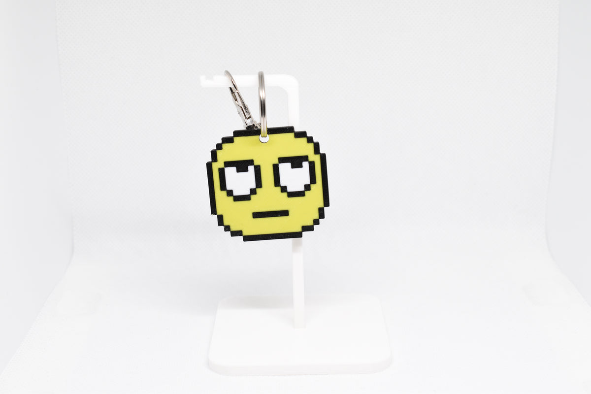 3D printed retro pixelated rolling eyes emoji keychain hanging on a white display stand by LayerPlay