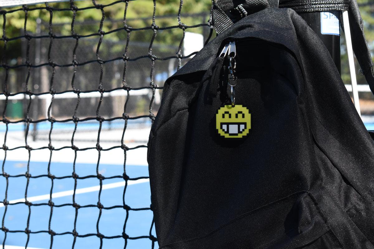 Pixelated smiling face emoji keychain clipped to a black backpack zipper outdoors near a tennis court by LayerPlay