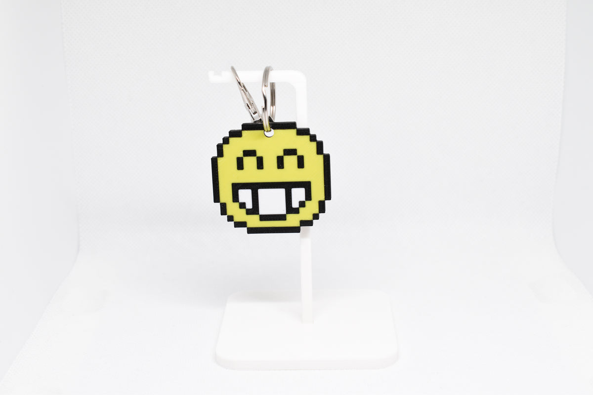 3D printed retro pixelated smiling face emoji keychain hanging on a white display stand by LayerPlay
