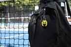 Pixelated squinting face emoji keychain clipped to a black backpack zipper outdoors near a tennis court by LayerPlay