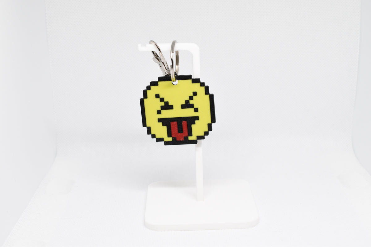 3D printed retro pixelated squinting face emoji keychain hanging on a white display stand by LayerPlay
