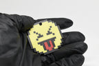 Hand in black glove holding 3D printed pixel squinting face emoji keychain front view detail by LayerPlay