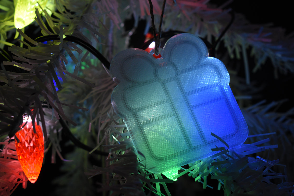 Translucent 3D printed holiday present ornament hanging on a white Christmas tree catching blue and green illuminated light.