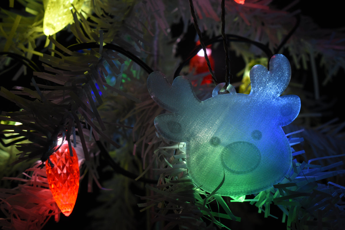 3D printed translucent reindeer ornament hanging on a white Christmas tree glowing with colorful tree light illumination.