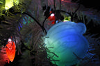 Translucent 3D printed Santa Claus ornament hanging on a white Christmas tree illuminated by glowing colorful holiday lights.