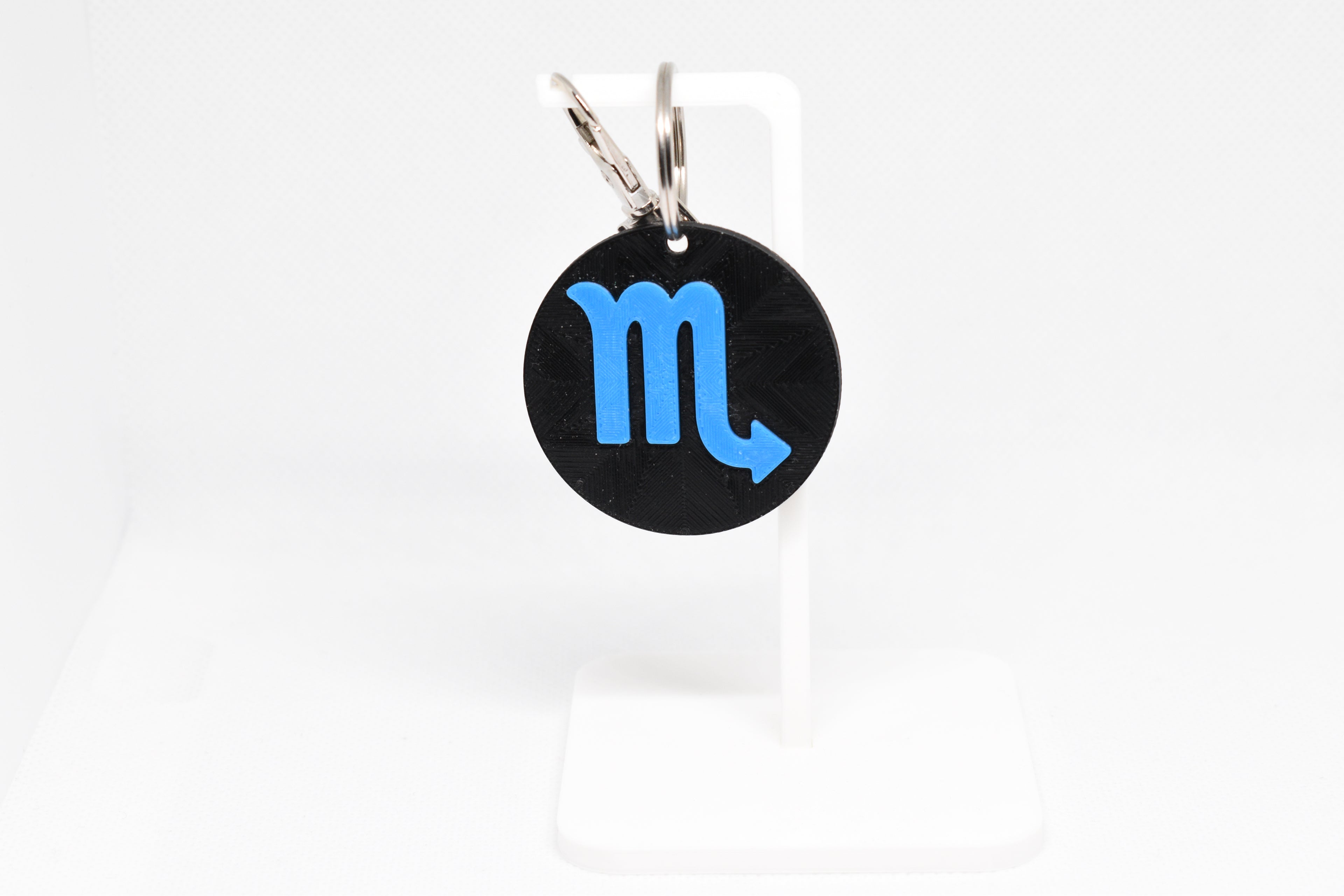 Scorpio zodiac keychain with blue symbol displayed on a white stand with metal ring and clip.