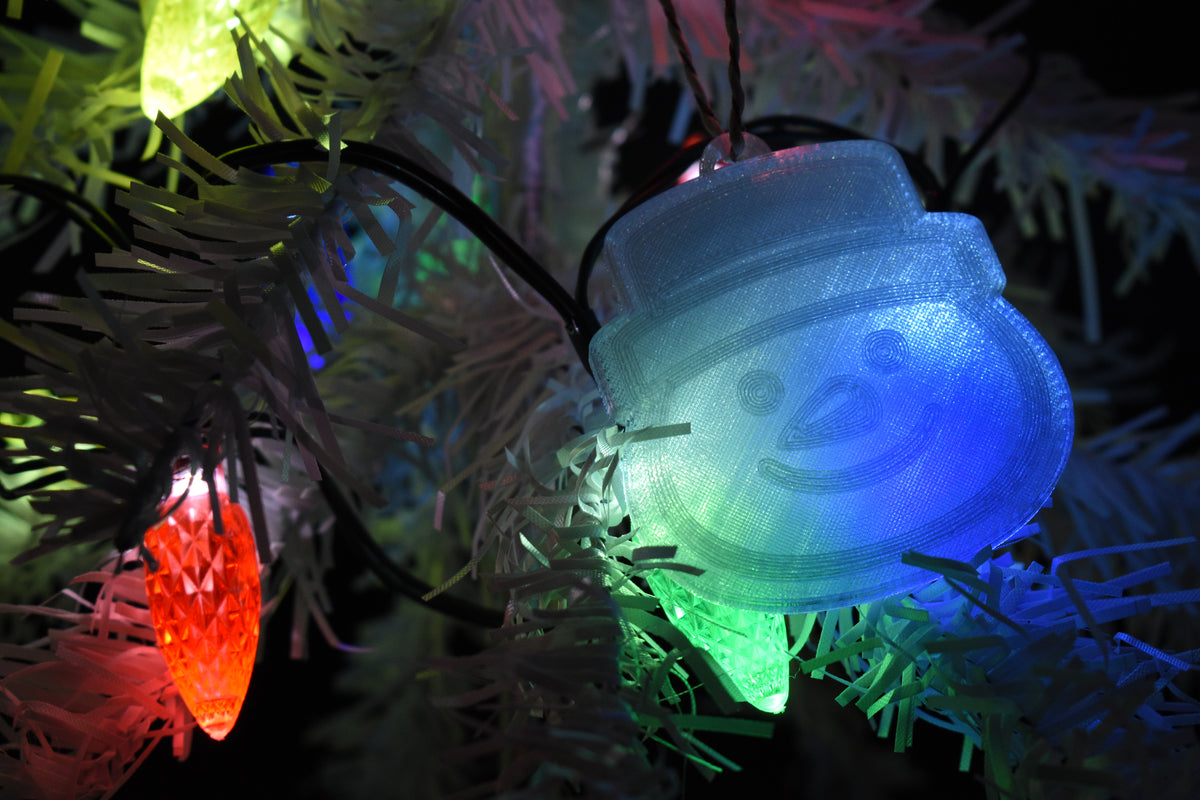 3D printed translucent snowman ornament with top hat hanging on a white Christmas tree lit up by colorful holiday string lights.