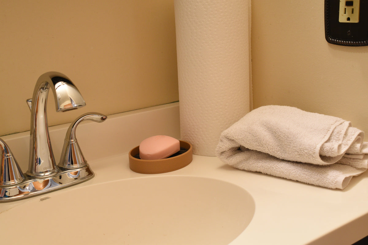 3D printed tan and black FlowGrid soap dish holding a pink soap bar on a bathroom vanity counter by LayerPlay Solara