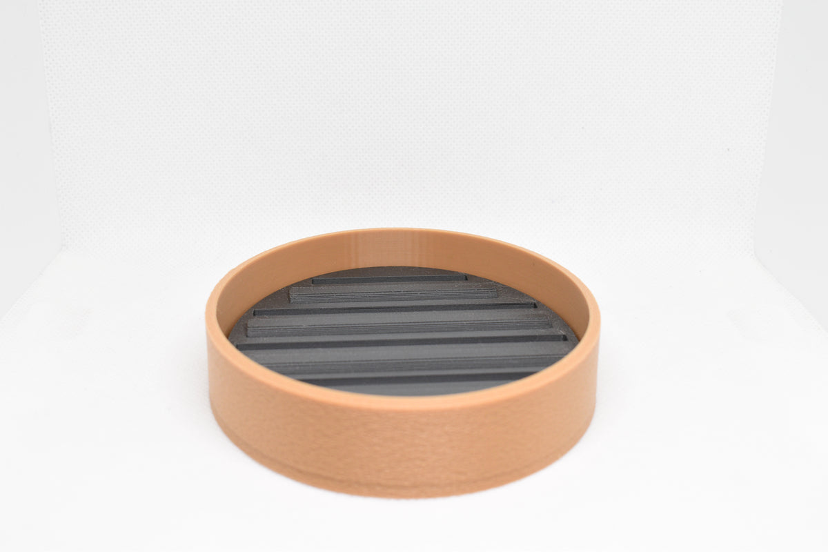 Low front studio view showing textured tan outer rim and black slotted grate insert of soap dish by LayerPlay Solara