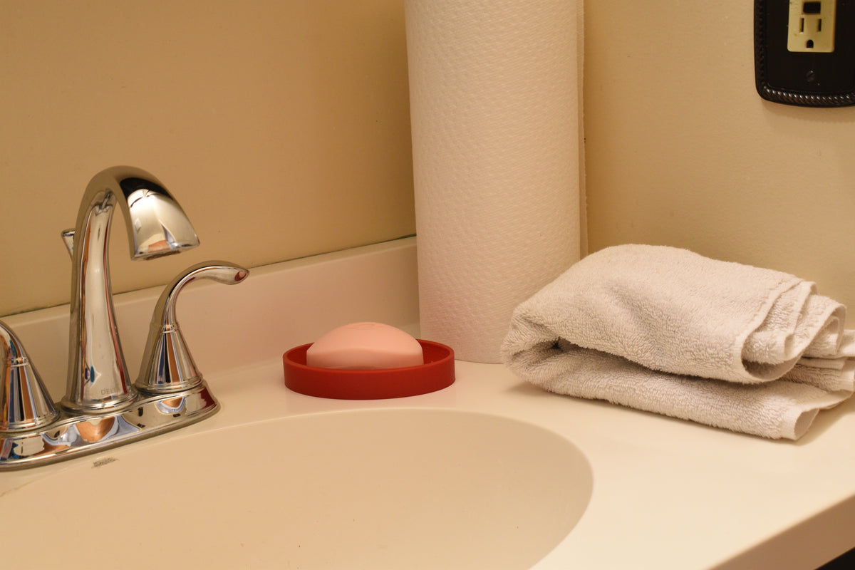 3D printed red TriLift soap dish holding a pink bar of soap on a bathroom vanity counter next to a chrome faucet by LayerPlay Solara