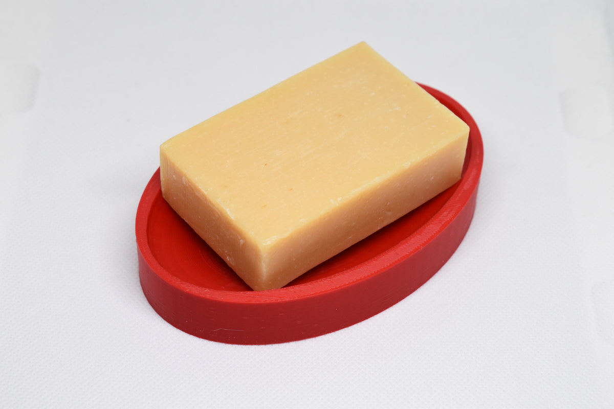 Natural rectangular bar soap resting on raised ridges of 3D printed red oval soap dish by LayerPlay Solara