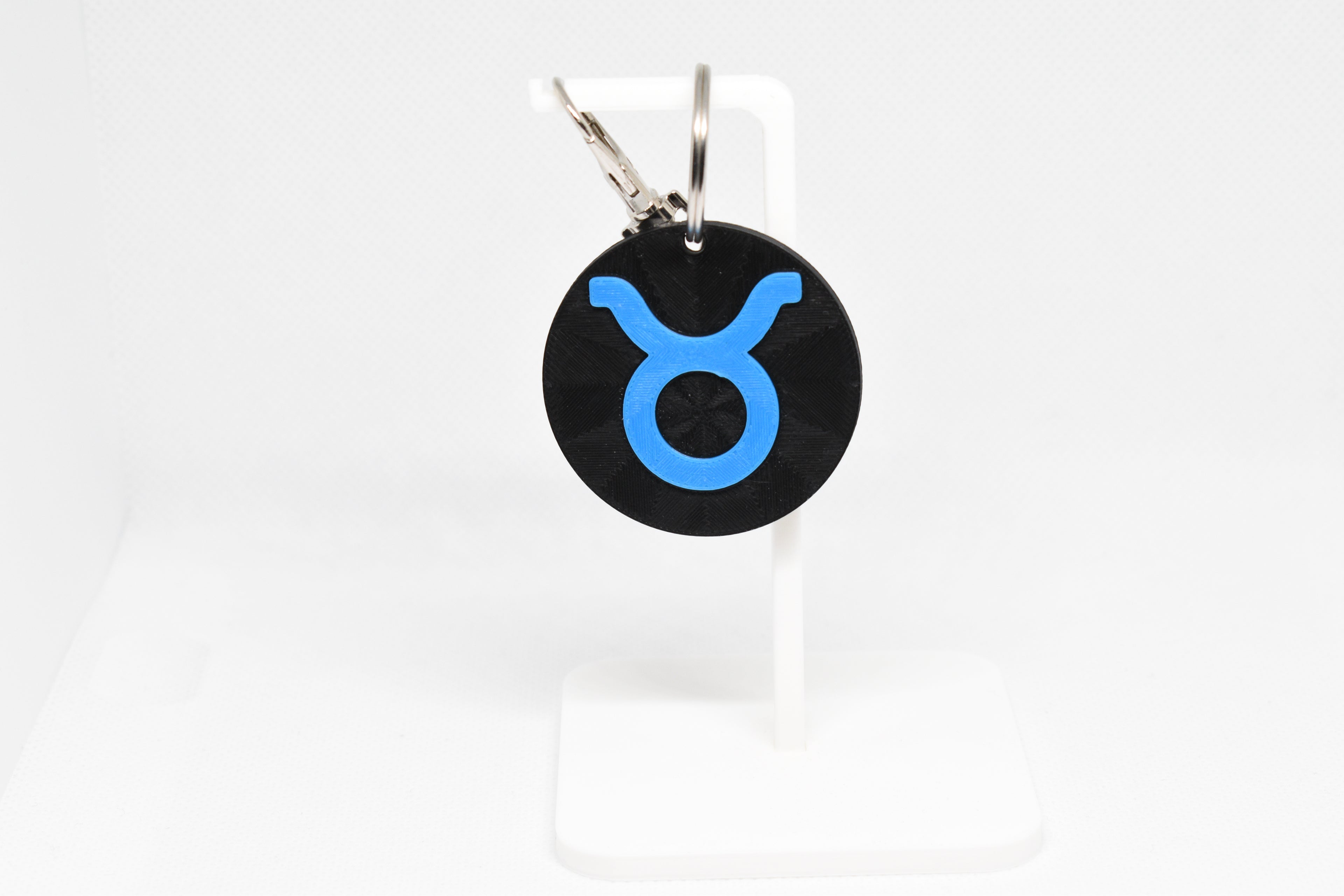 Taurus zodiac keychain with blue symbol displayed on a white stand with metal ring and clip.