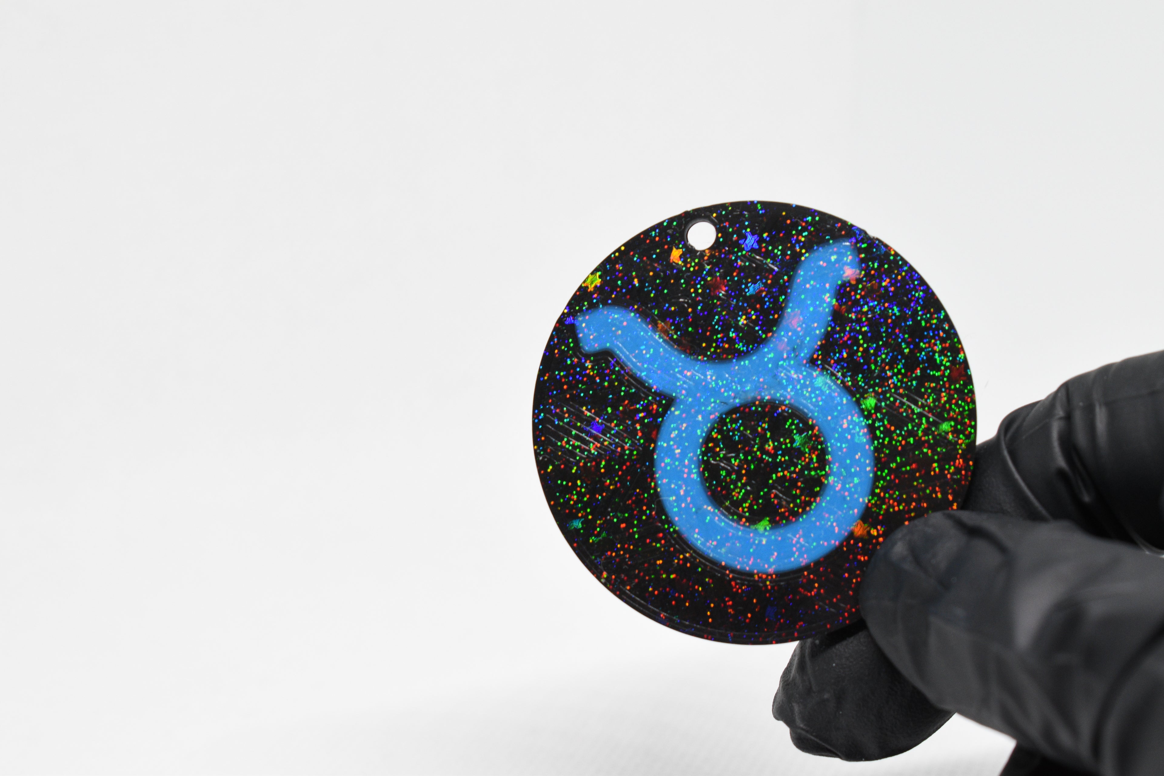 Taurus zodiac keychain with blue symbol and glitter‑black cosmic background held in a gloved hand.