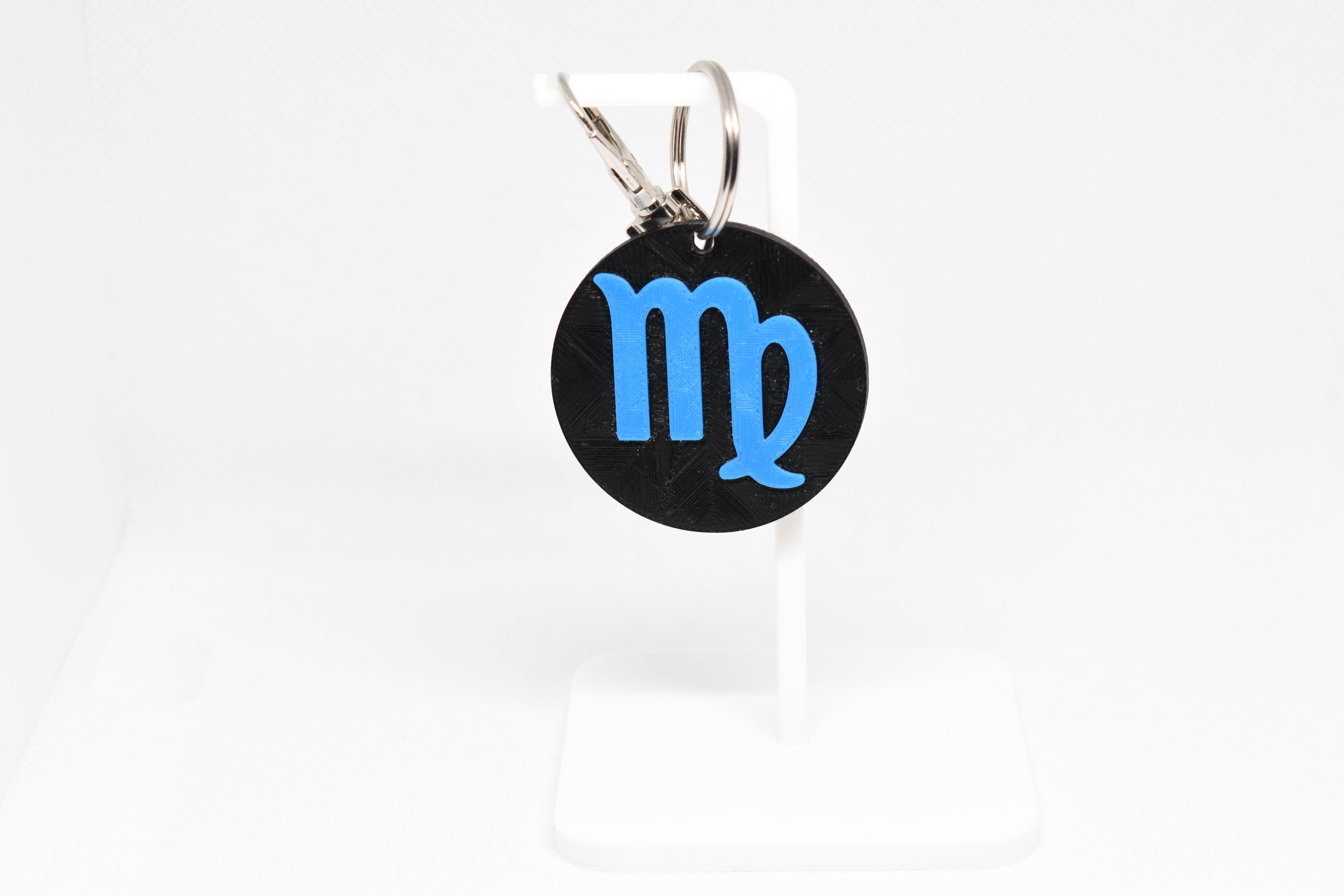 Virgo zodiac keychain with blue symbol displayed on a white stand with metal ring and clip.