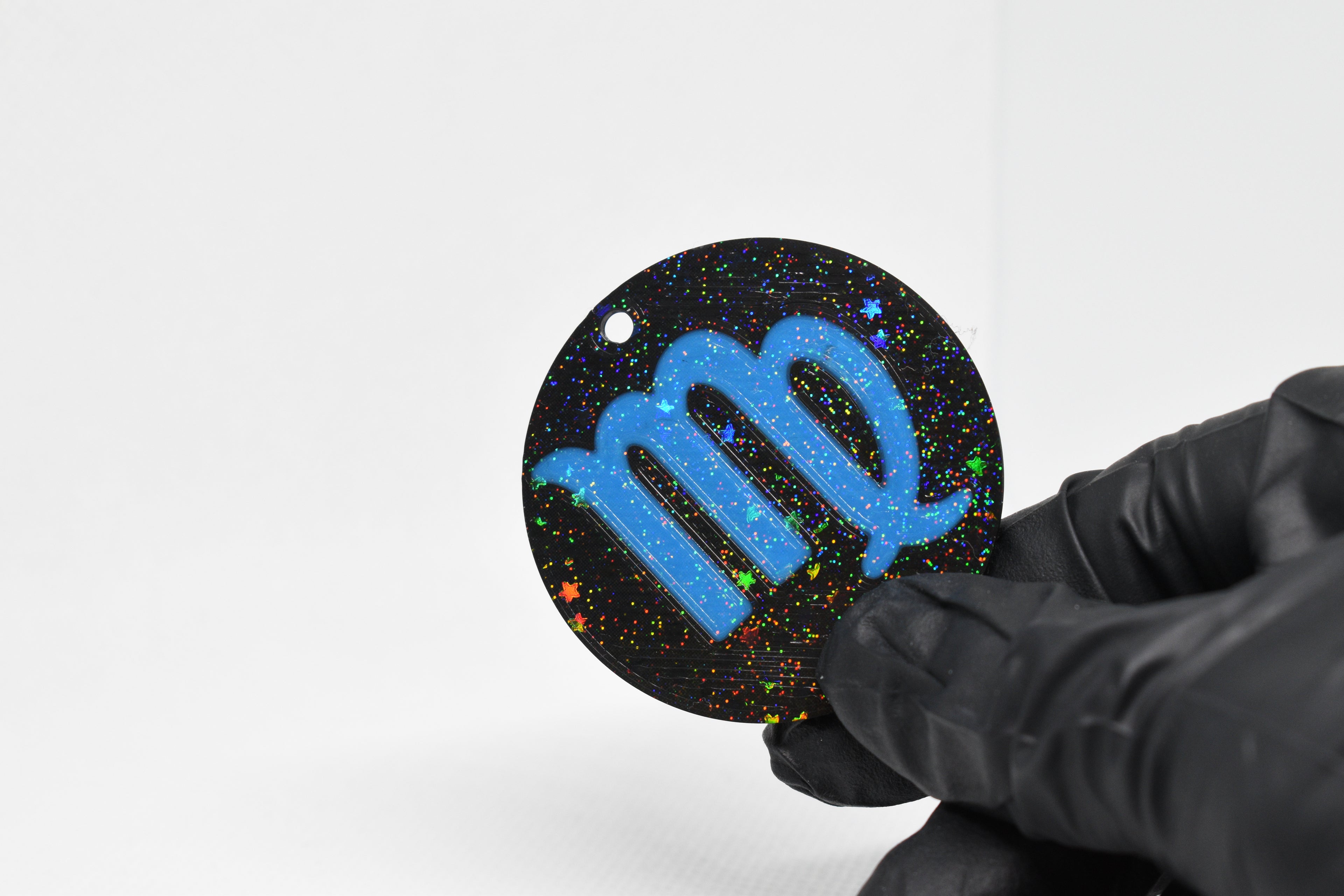 Virgo zodiac keychain with blue symbol and glitter‑black cosmic background held in a gloved hand.