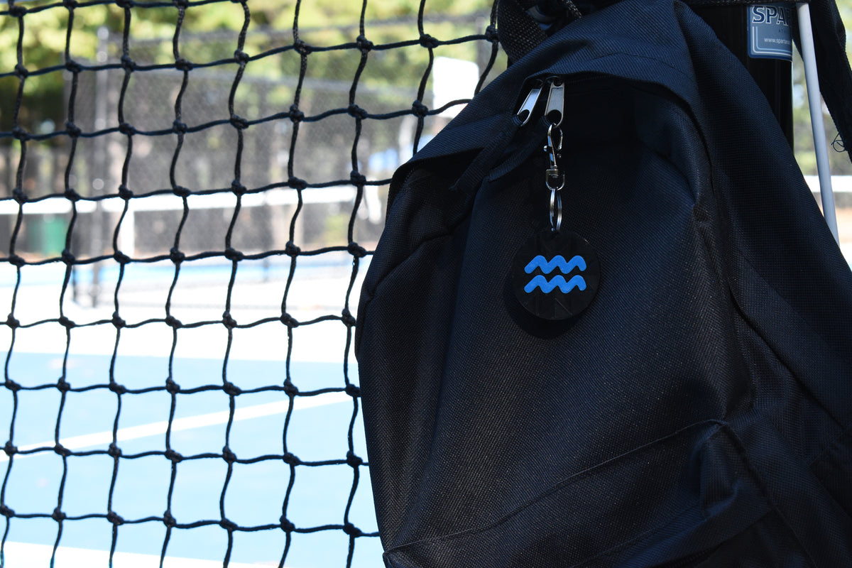 3D printed aquarius zodiac keychain clipped to a black backpack zipper outdoors near a tennis court by LayerPlay