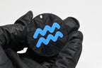 Hand in black glove holding 3D printed blue aquarius zodiac symbol keychain front view detail by LayerPlay