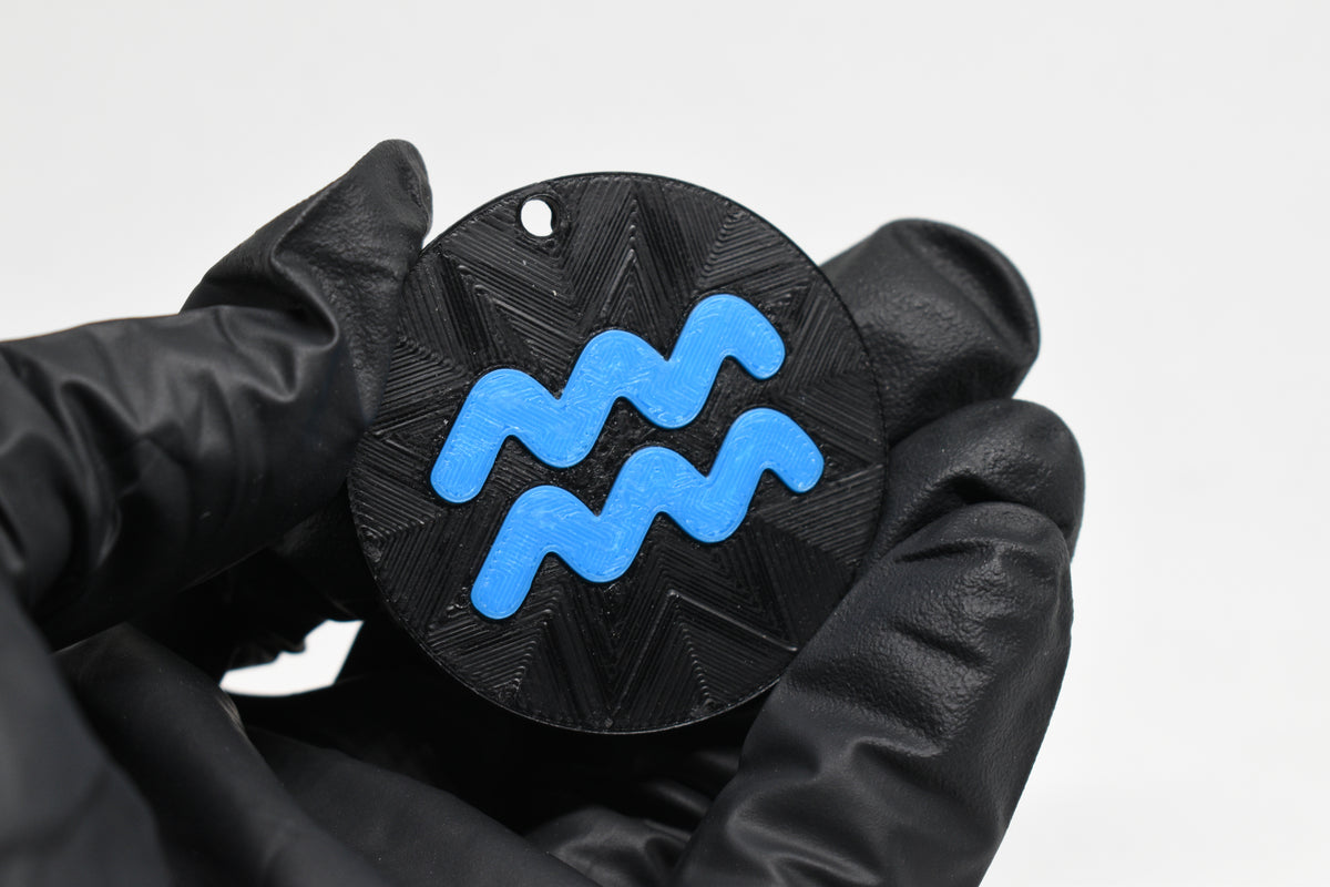 Hand in black glove holding 3D printed blue aquarius zodiac symbol keychain front view detail by LayerPlay