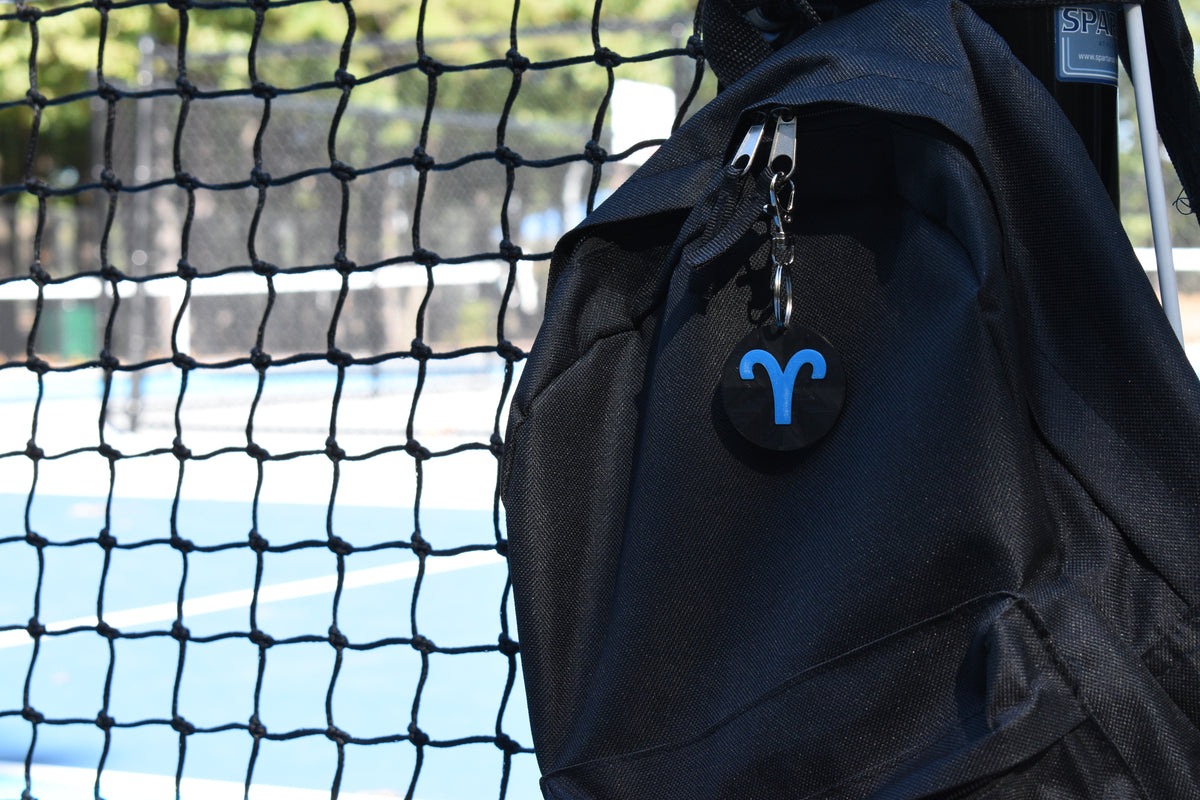 3D printed aries zodiac keychain clipped to a black backpack zipper outdoors near a tennis court by LayerPlay