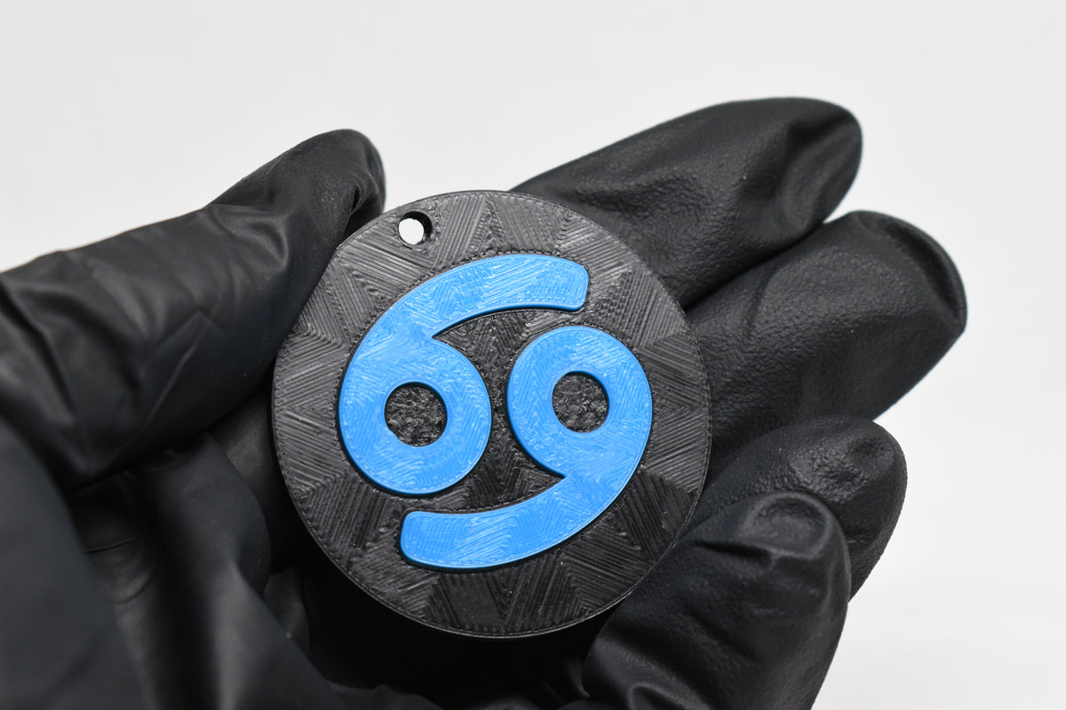 Hand in black glove holding 3D printed blue cancer zodiac symbol keychain front view detail by LayerPlay