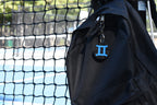 3D printed gemini zodiac keychain clipped to a black backpack zipper outdoors near a tennis court by LayerPlay