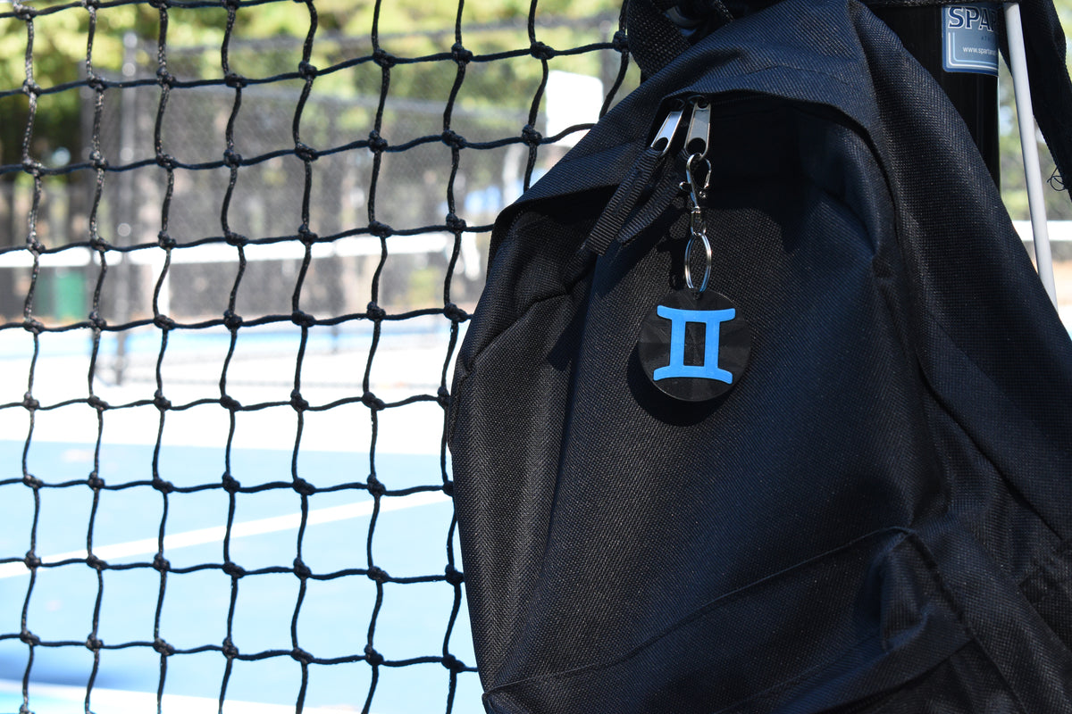 3D printed gemini zodiac keychain clipped to a black backpack zipper outdoors near a tennis court by LayerPlay