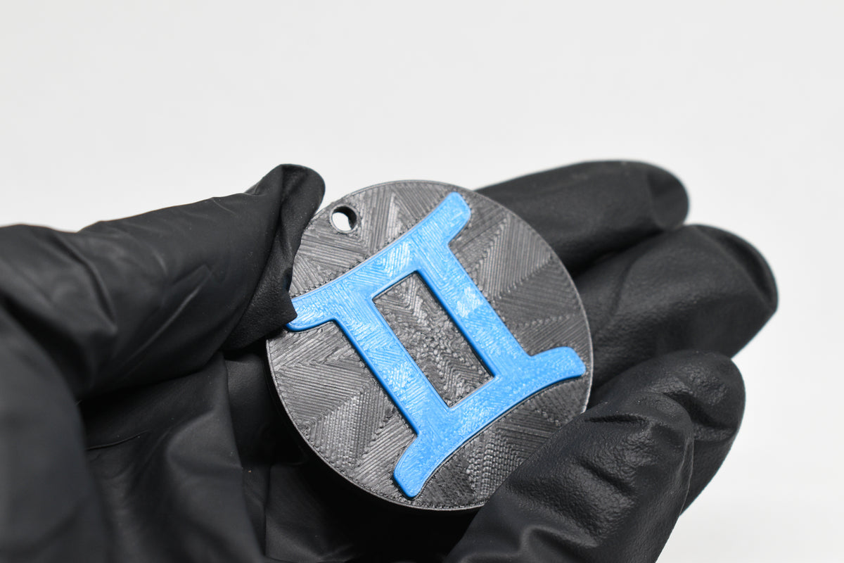 Hand in black glove holding 3D printed blue gemini zodiac symbol keychain front view detail by LayerPlay