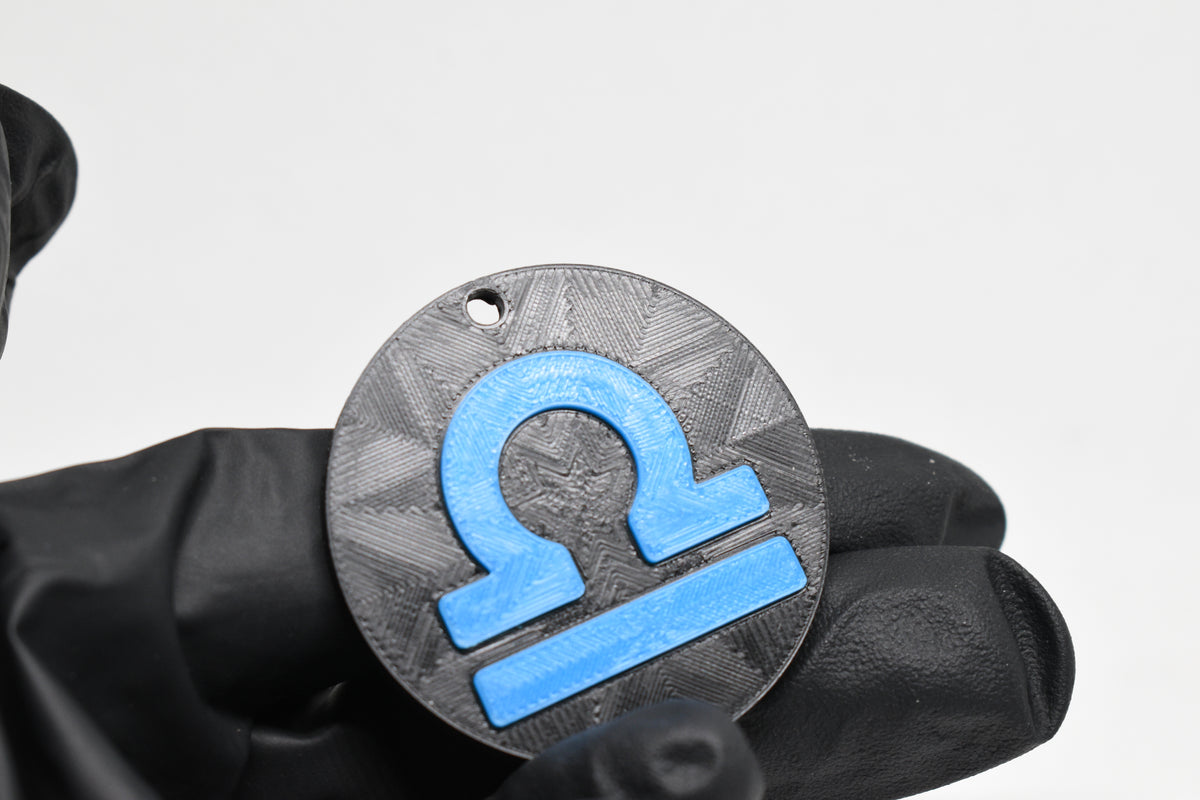 Hand in black glove holding 3D printed blue libra zodiac symbol keychain front view detail by LayerPlay