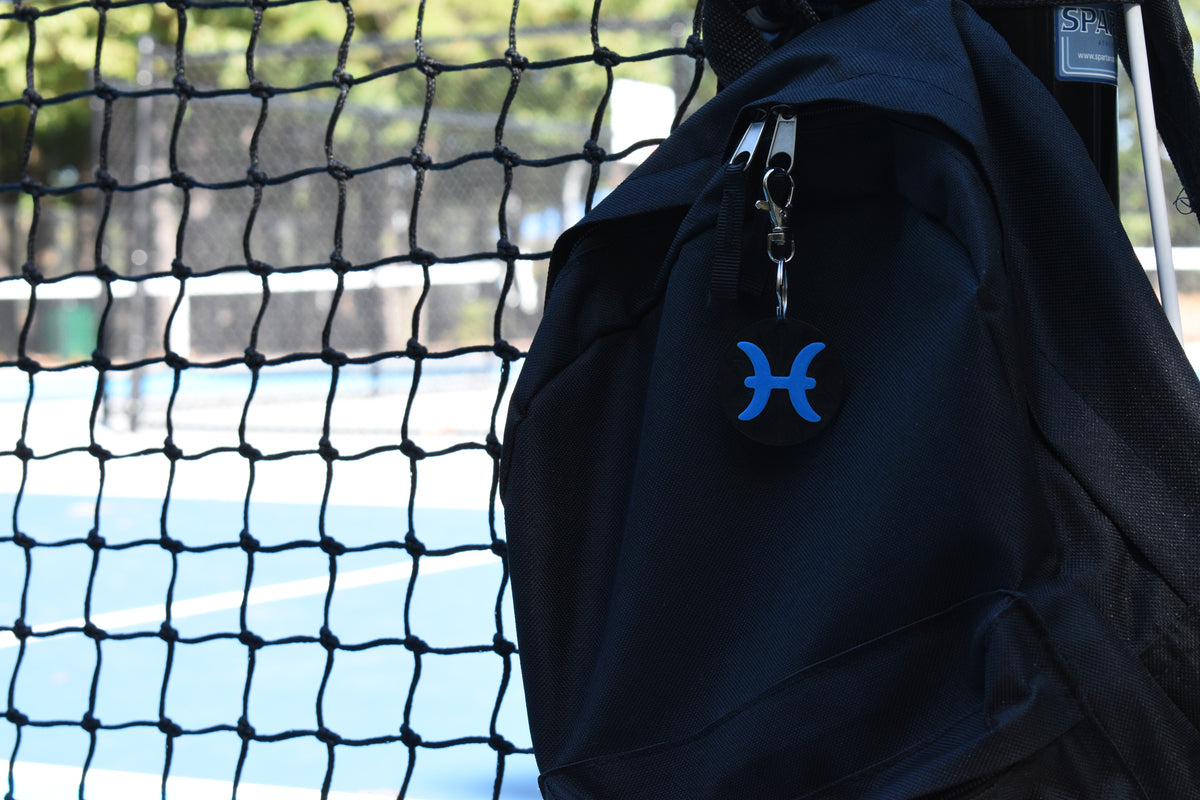 3D printed pisces zodiac keychain clipped to a black backpack zipper outdoors near a tennis court by LayerPlay