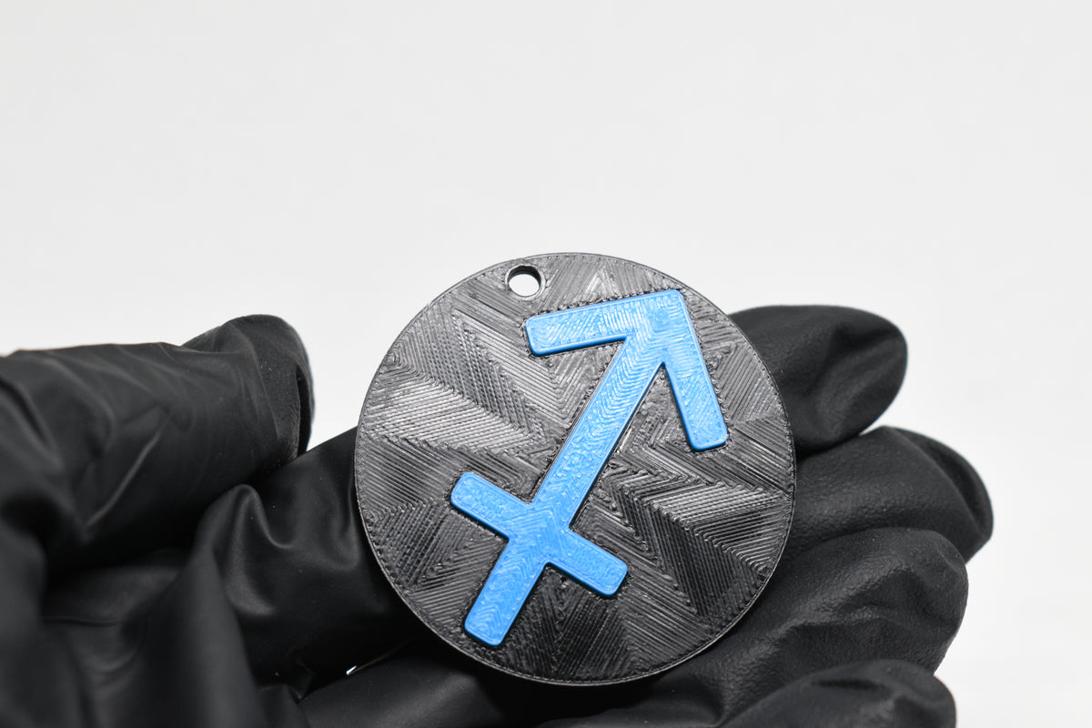 Hand in black glove holding 3D printed blue sagittarius zodiac archer symbol keychain front view detail by LayerPlay