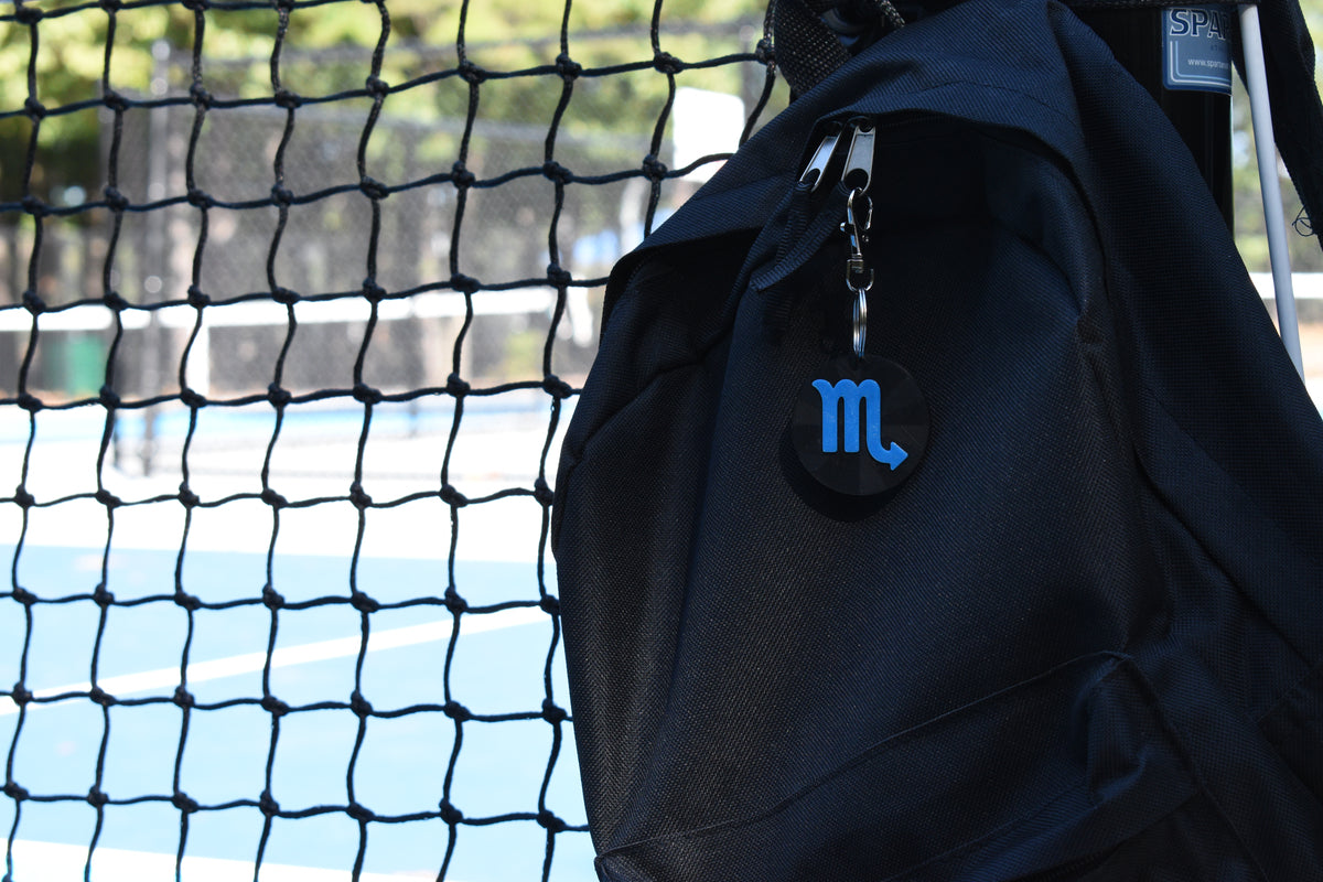 3D printed scorpio zodiac keychain clipped to a black backpack zipper outdoors near a tennis court by LayerPlay
