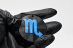 Hand in black glove holding 3D printed blue scorpio zodiac symbol keychain front view detail by LayerPlay